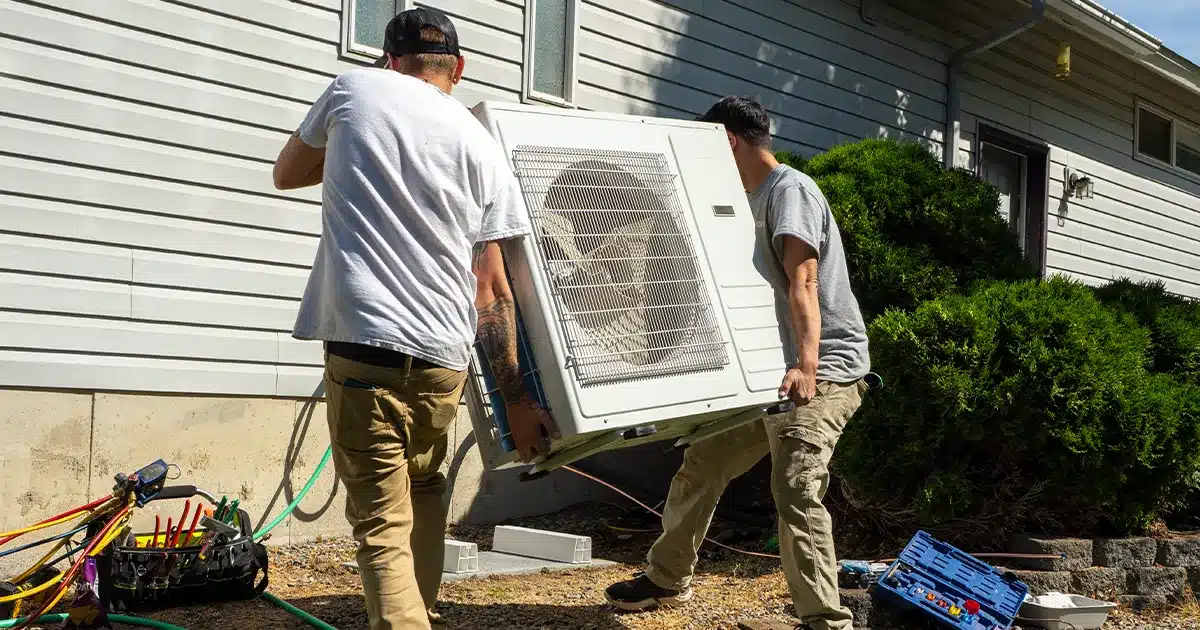 Two HVAC installers carry a heat pump towards its final installation location